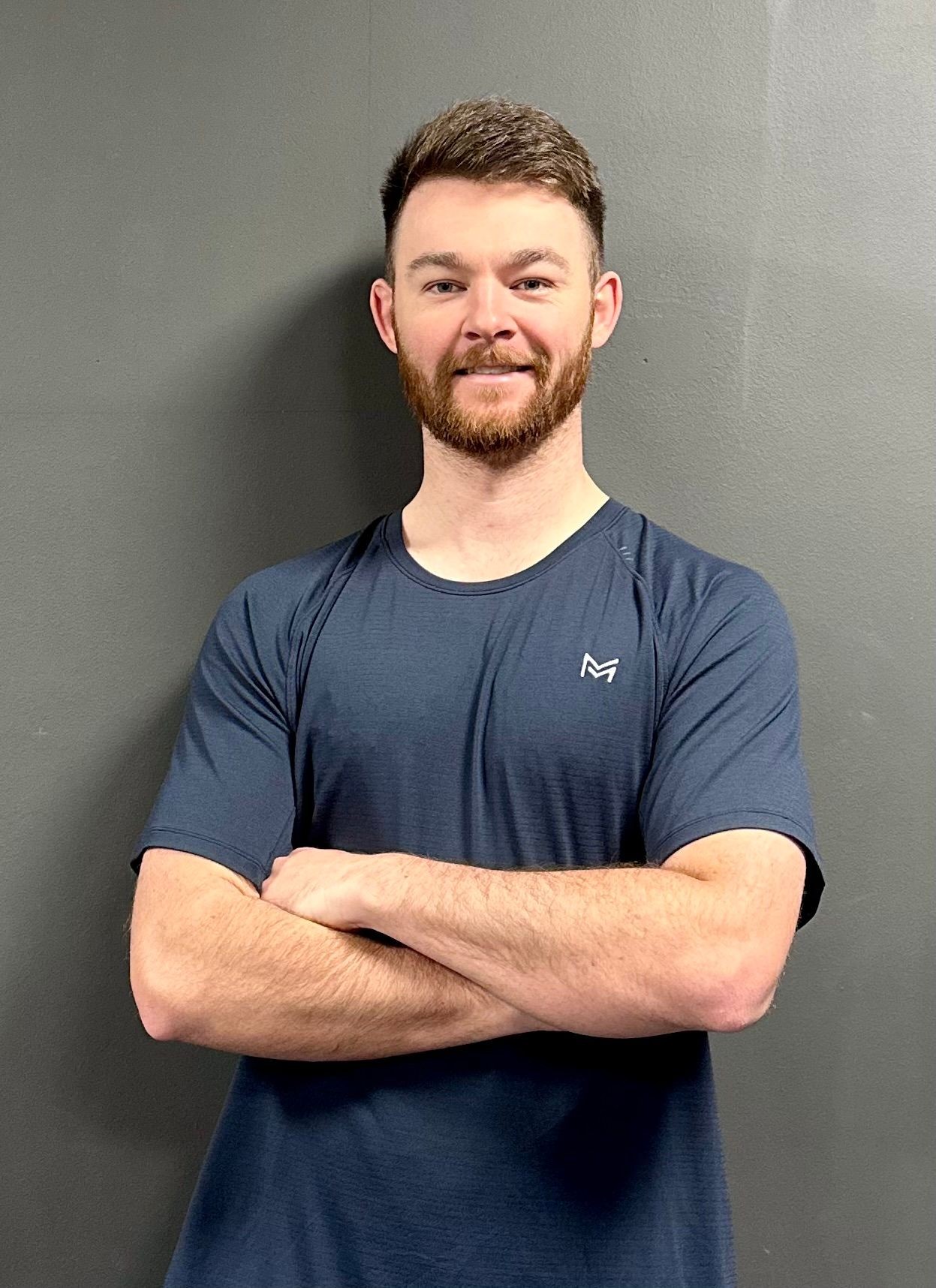 Man with a beard wearing a blue shirt, standing with arms crossed against a gray background.