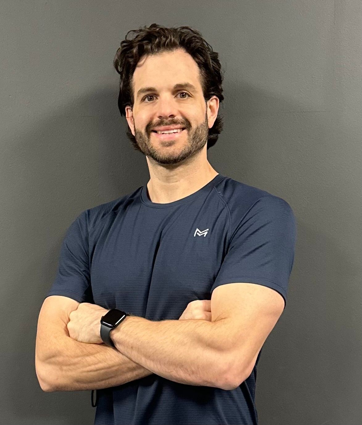 Man in navy shirt with smartwatch, smiling and crossing arms against gray wall.